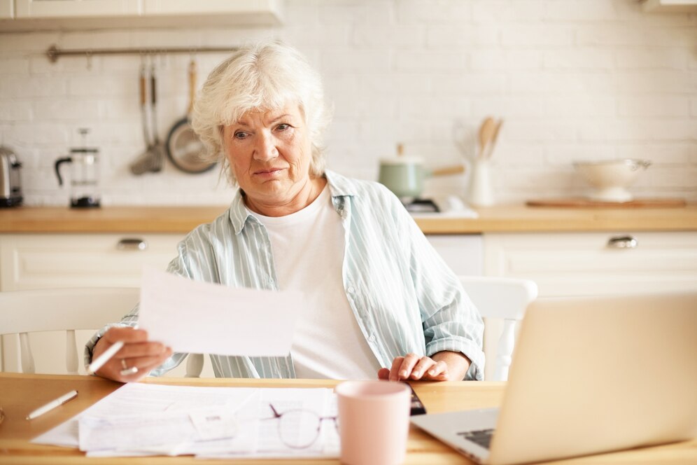elderly-housewife-with-gray-hair-sitting-kitchen-with-open-laptop-papers-table-having-emotional-frustrated-facial-expression-shocked-with-debt-amount-while-paying-dome